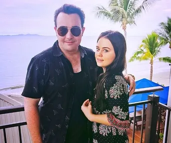 A man in sunglasses and a woman in a patterned dress pose on a balcony with a beach and palm trees in the background.
