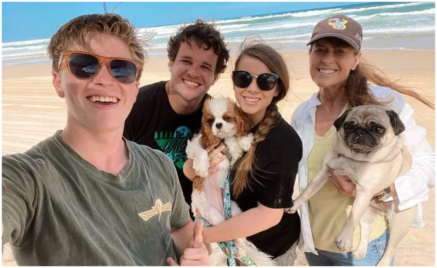 Family smiling on a beach holding two dogs, with the ocean in the background.