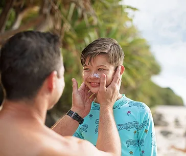 Parent applying sunscreen to a smiling child on a beach, surrounded by green trees.