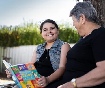 Child and older woman reading "Favorite Tales" book together outdoors, both smiling and enjoying the moment.