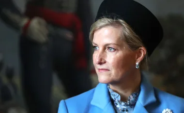 A woman in a blue coat with short blonde hair and a black hat, looking to the right, set against a blurred background.