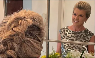 A woman with a braided hairstyle looks at her reflection in a mirror, wearing a shiny, patterned top.