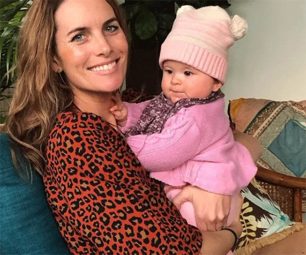 Woman in a leopard print top holding a baby in a pink outfit and hat, both smiling, seated on a colorful couch.