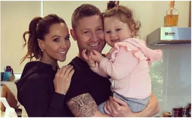 A family posing in a kitchen; two adults smile while holding a child, showing a warm and happy moment together.