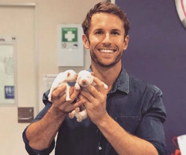 Man smiling while holding two small puppies indoors.