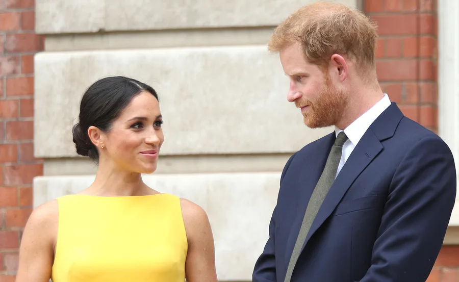 A woman in a yellow dress and a man in a suit smile at each other outside a brick building.