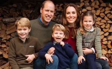 A family of five smiling, sitting in front of stacked firewood.