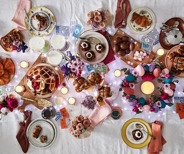 Festive table with hot cross buns, pastries, flowers, candles, and decorative elements arranged colorfully.