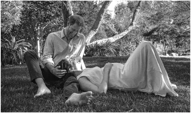 A couple sits on the grass laughing, with trees in the background.