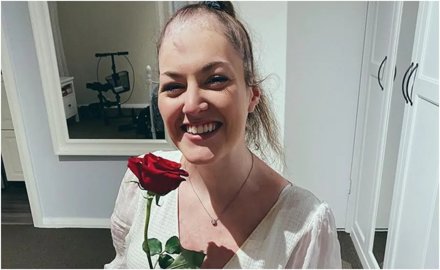 A woman smiling, holding a red rose in a room with a mirror and exercise equipment in the background.
