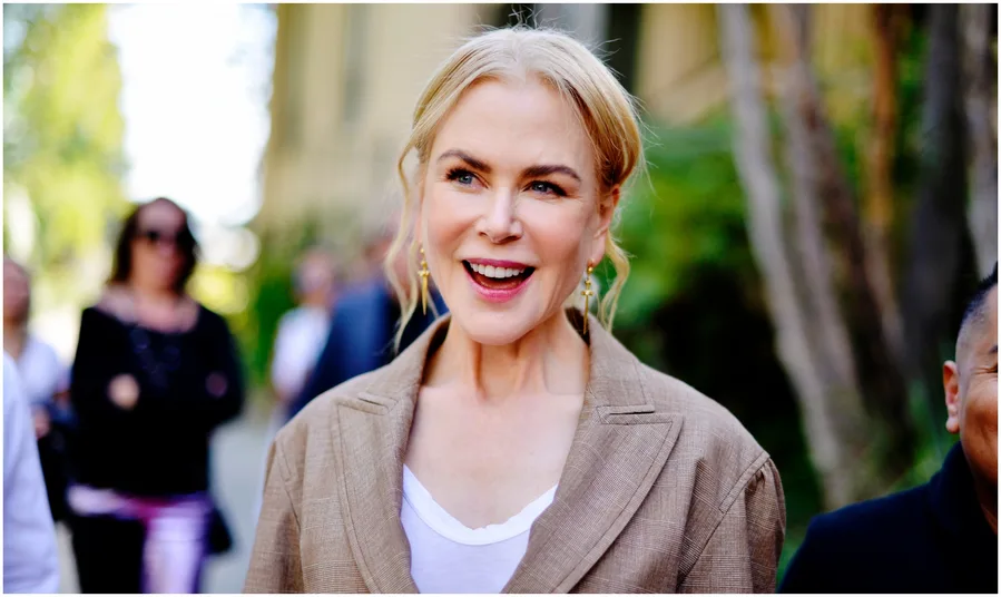 A smiling woman with light hair in a bun, wearing a brown blazer and white top, outdoors with blurred people in the background.