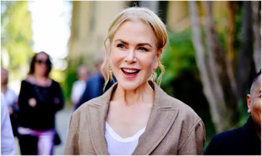 A smiling woman with light hair in a bun, wearing a brown blazer and white top, outdoors with blurred people in the background.