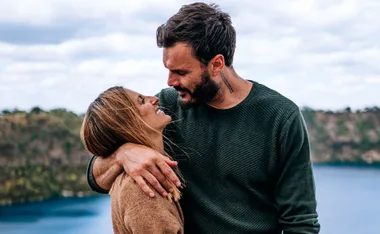 A couple smiling at each other outdoors with a scenic backdrop of water and hills.