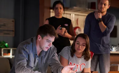 Group of four people in a dimly lit room, looking intently at a laptop screen, appearing concerned.