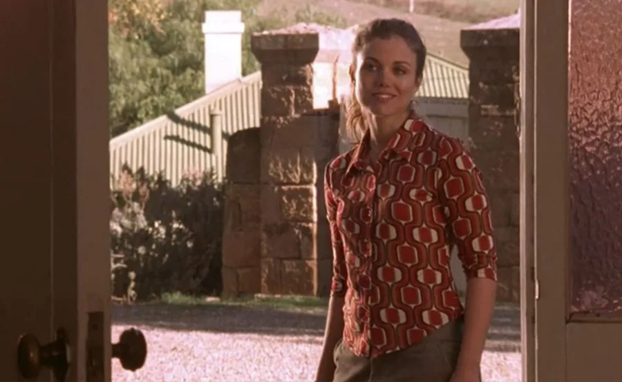 A woman in a patterned top stands in a doorway, with a stone wall and greenery visible outside.