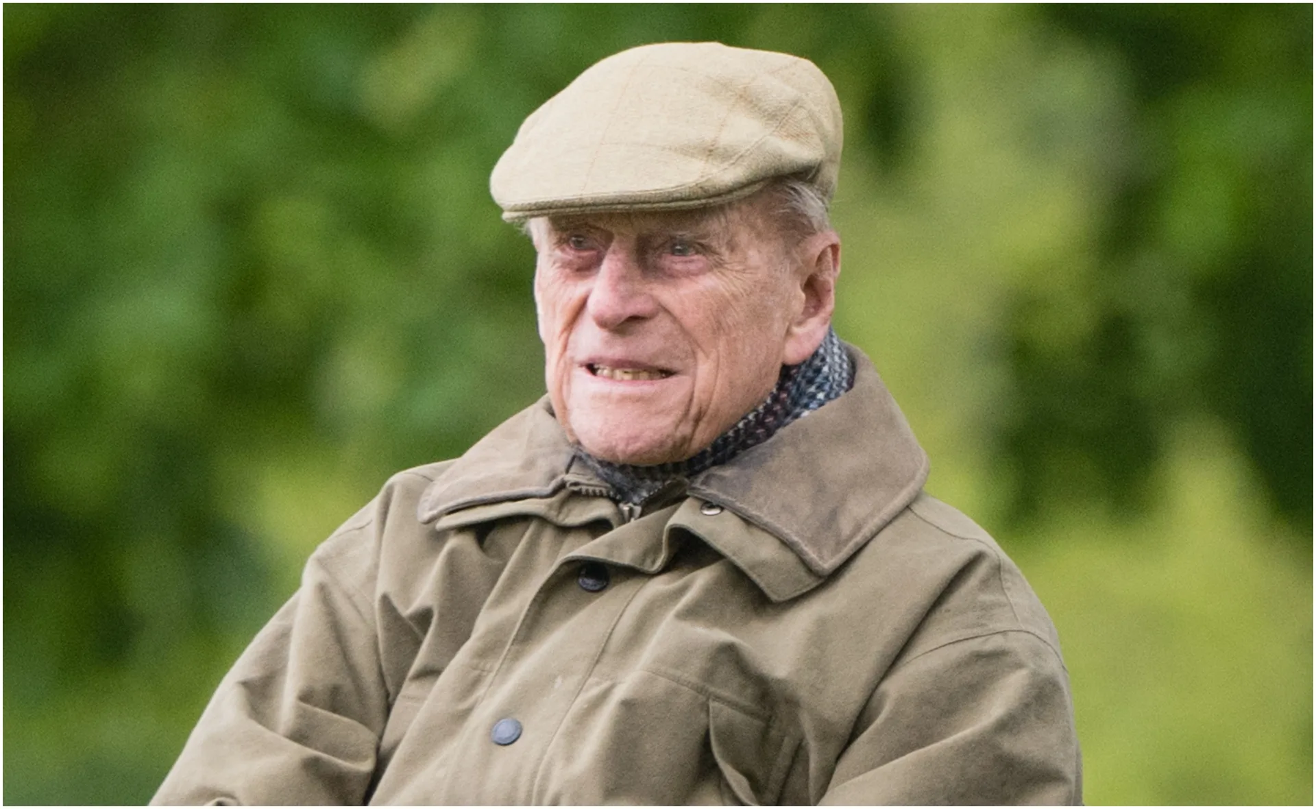 Elderly man in a flat cap and coat, sitting outdoors with a neutral expression.