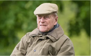 Elderly man in a flat cap and coat, sitting outdoors with a neutral expression.