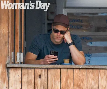 Man in sunglasses and cap gazing at phone, leaning on a wooden counter, with "Woman's Day" text above.