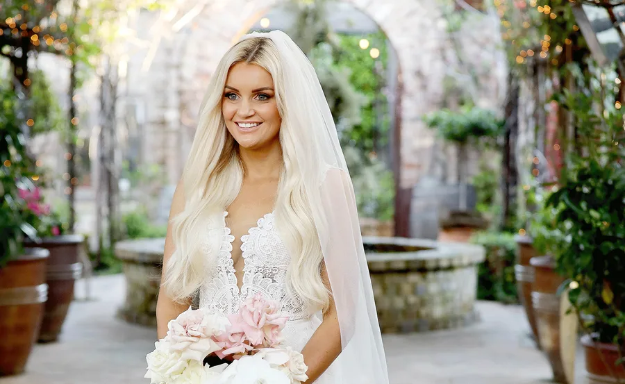 Bride in a lace wedding dress and veil, holding a bouquet, smiling in a garden setting.