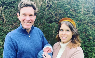 A couple smiles outside, holding a newborn baby wrapped in a blue blanket.