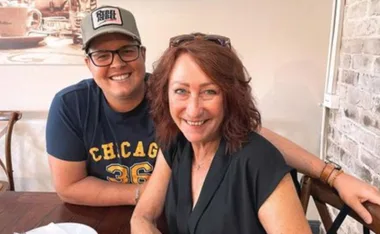 Two people smiling at a cafe table, one wearing glasses and a cap, the other with auburn hair and sunglasses on head.