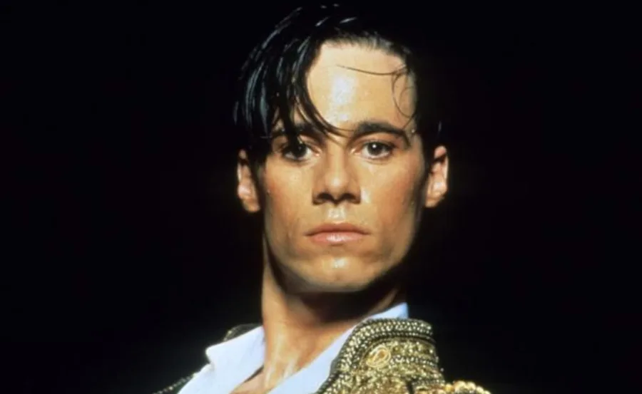 A person with wet hair and intense expression wearing an ornate gold-and-black costume against a dark background.
