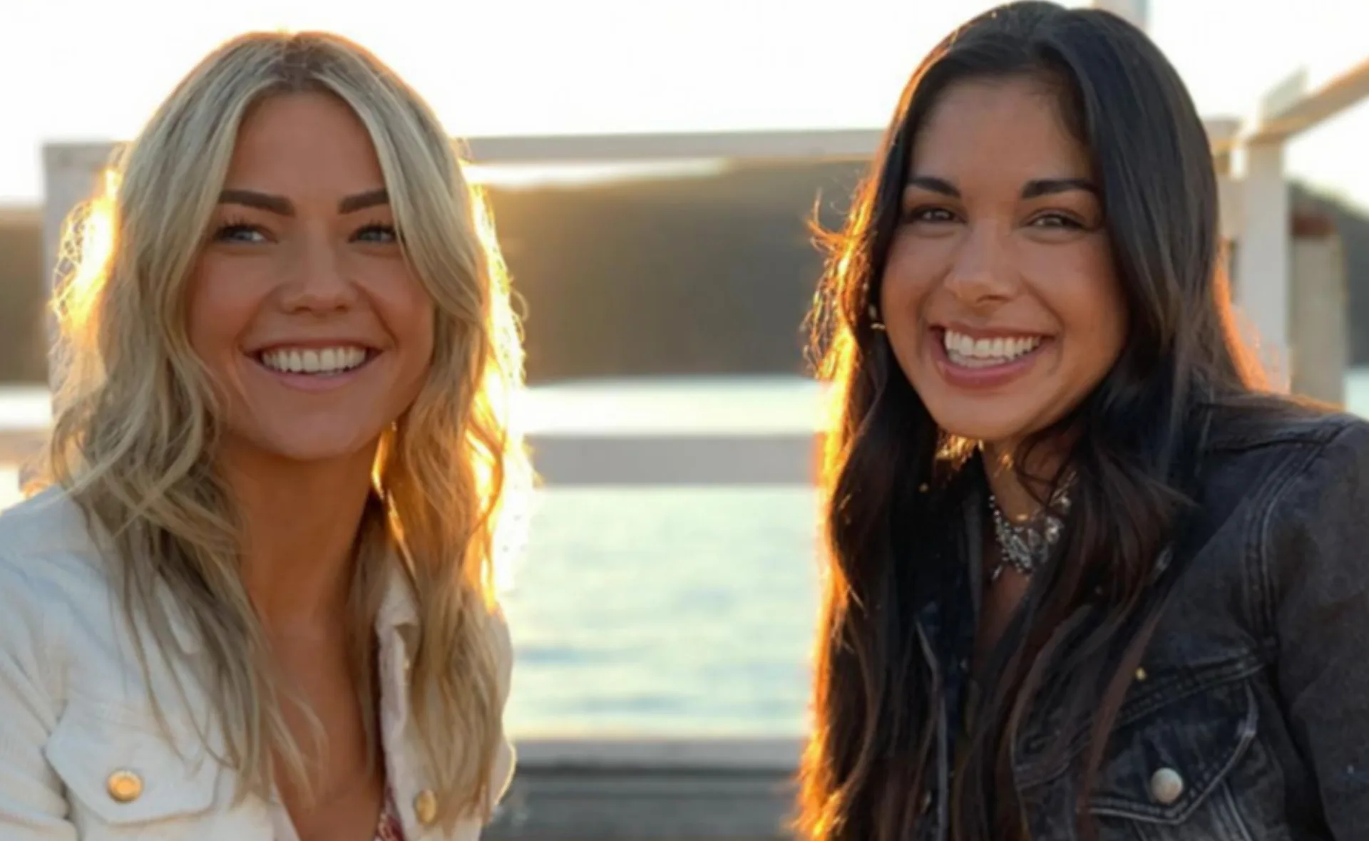 Two women smiling outdoors in sunlight, one with blonde hair and the other with dark hair, both wearing jackets.