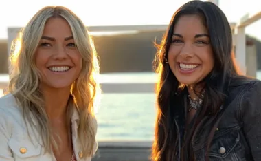 Two women smiling outdoors in sunlight, one with blonde hair and the other with dark hair, both wearing jackets.