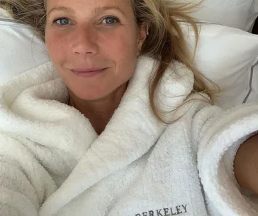 Person in a fluffy white robe with "Berkeley" embroidered, lying on a bed, looking at the camera.
