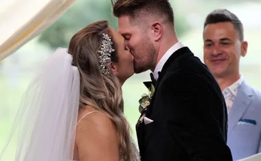 A bride and groom share a kiss at their wedding ceremony, with an officiant smiling in the background.