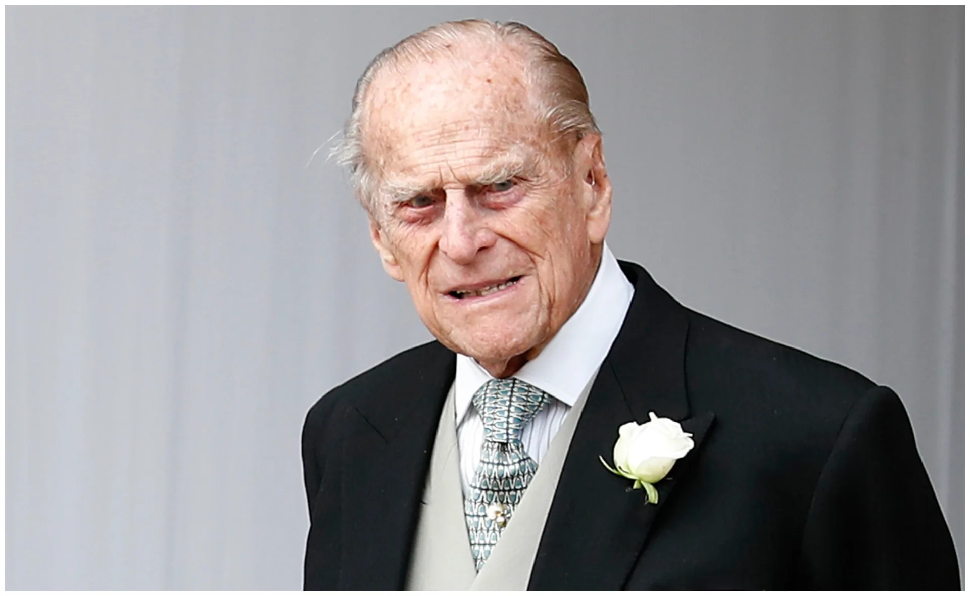 Man in a black suit with a white rose boutonniere and patterned tie looking ahead.