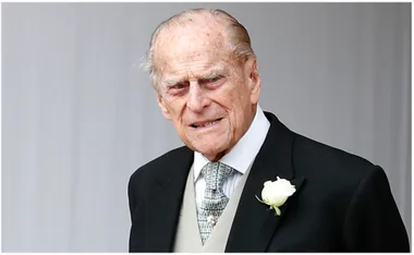Man in a black suit with a white rose boutonniere and patterned tie looking ahead.