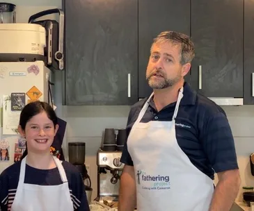 Father and daughter wearing aprons in a kitchen, smiling at the camera; part of the Fathering Project initiative.