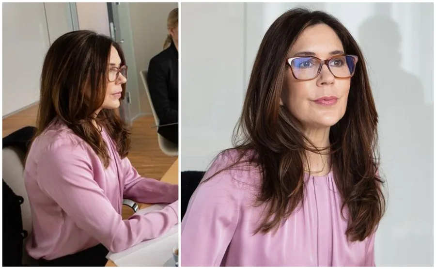A woman with long hair and glasses wearing a pink shirt, seated at a table in an office setting.