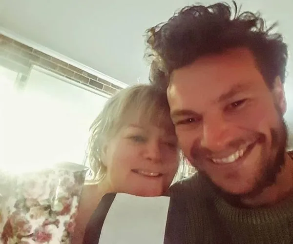 Two people smiling for a close-up selfie indoors, one holding a floral mug.