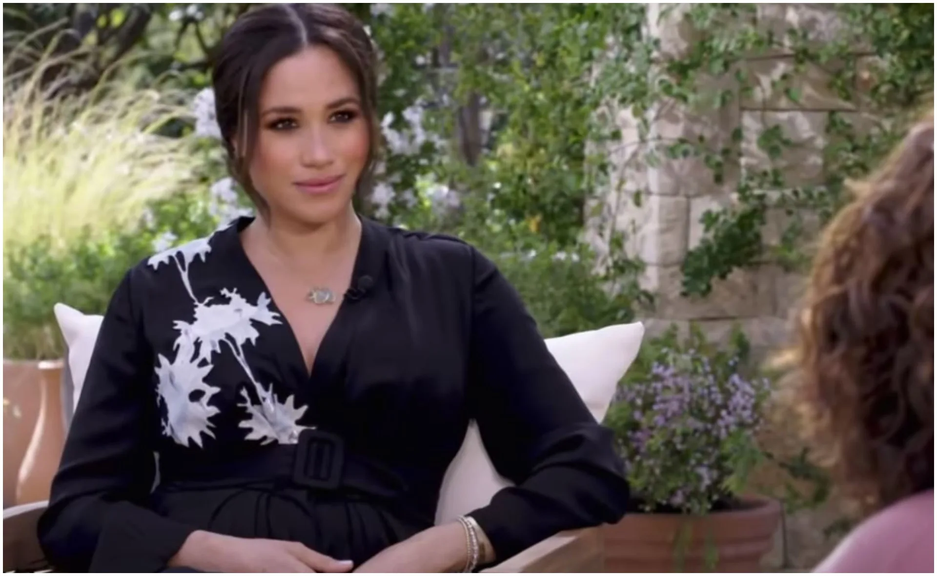 Woman in black dress with white floral design sitting outdoors, speaking in an interview setting.