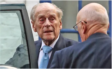 Elderly man in a suit near a car, speaking with another man, appearing concerned or serious.