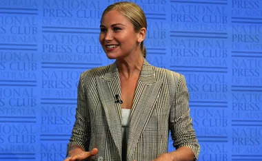 A woman in a checkered blazer speaks at the National Press Club of Australia, smiling with a microphone attached.