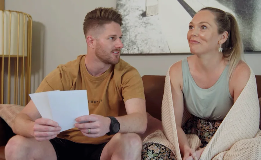 Man and woman sitting on a couch, man holding papers, both looking at each other pleasantly.