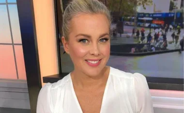Smiling person with blonde hair in a white top, sitting indoors with a window and blurred outdoor view behind.