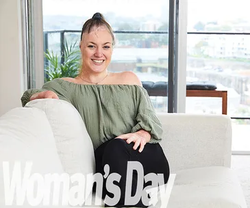 Woman smiling on a sofa in an apartment with city view, wearing an off-shoulder top. "Woman's Day" logo at the bottom.