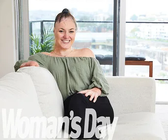 Woman smiling on a sofa in an apartment with city view, wearing an off-shoulder top. "Woman's Day" logo at the bottom.