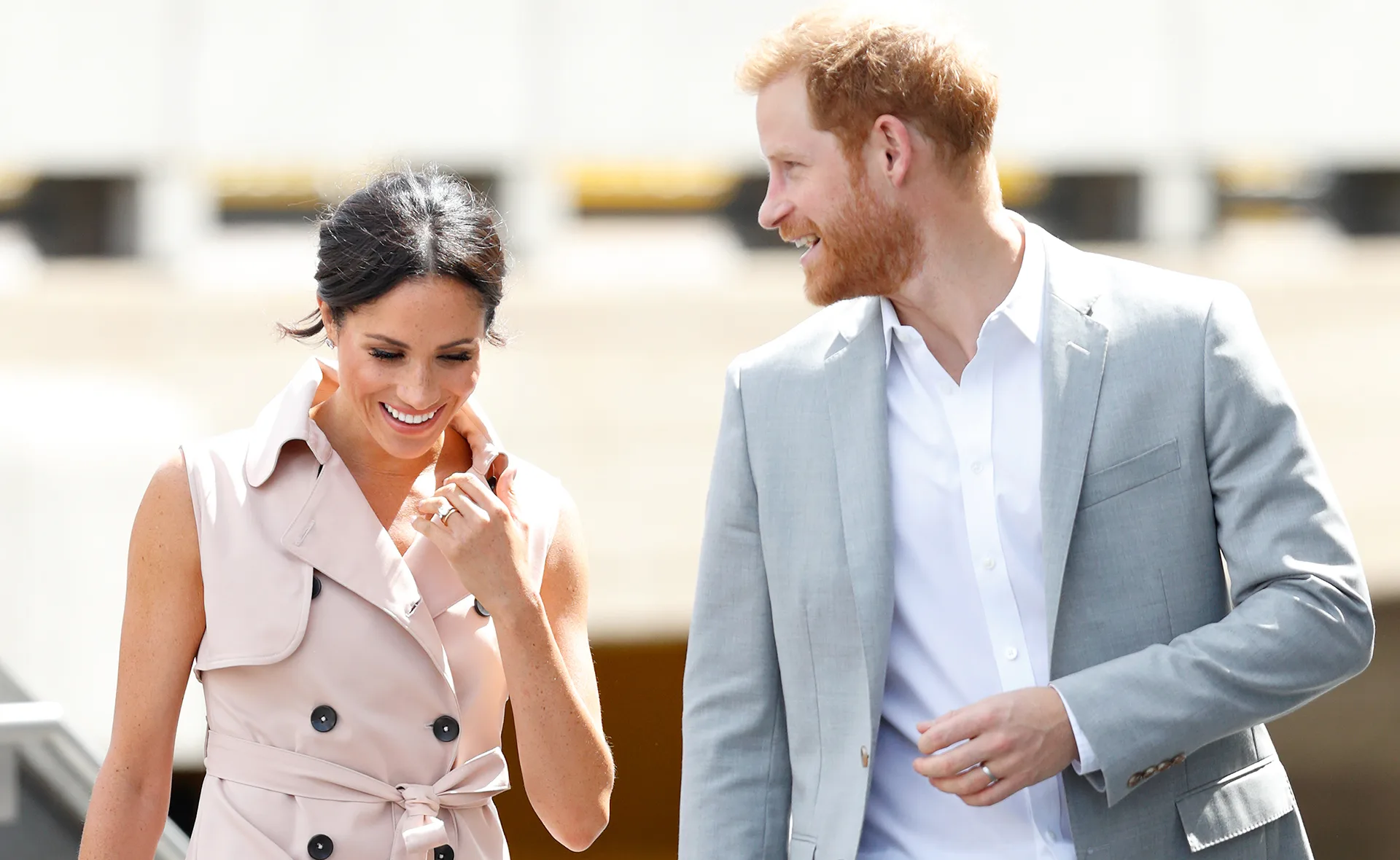 A smiling couple walking together, one wearing a light coat and the other a light suit, under the bright sun.