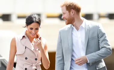 A smiling couple walking together, one wearing a light coat and the other a light suit, under the bright sun.