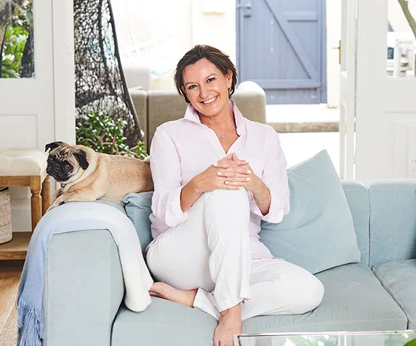 Woman in a white outfit sitting on a blue couch with a pug, smiling in a bright, cozy living room.