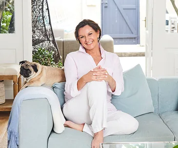 Woman in a white outfit sitting on a blue couch with a pug, smiling in a bright, cozy living room.