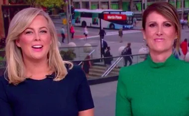 Two news anchors smiling with a city street and a bus in the background.