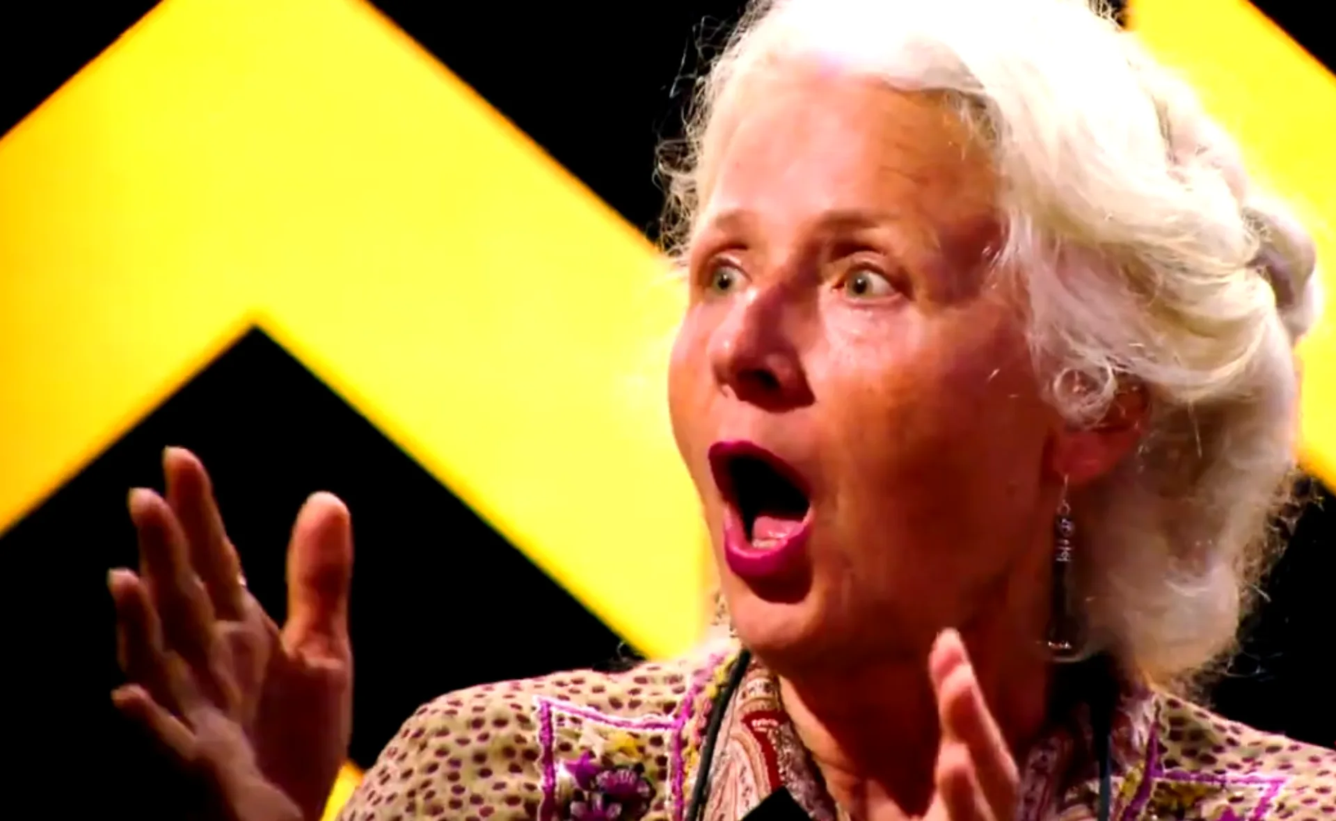 A surprised elderly woman in patterned clothes and earrings, with a yellow zigzag backdrop.