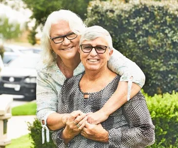 Two elderly women, both with short white hair and glasses, smiling warmly while embracing outdoors.