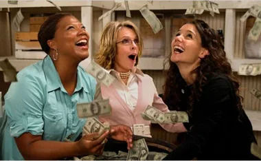 Three women look up joyfully as money falls around them indoors.